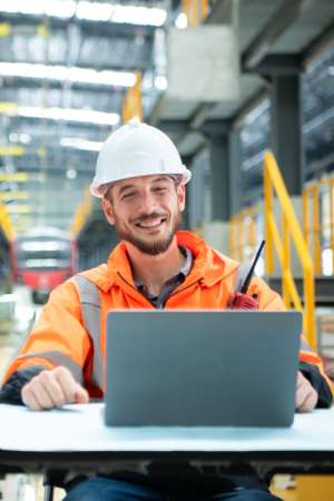 Un giovane tecnico o ingegnere con barba e casco protettivo bianco sorride seduto a un tavolo da lavoro davanti a un computer portatile aperto, indossando una giacca da lavoro ad alta visibilit&agrave; arancione e grigia con dettagli catarifrangenti e una radio ricetrasmittente agganciata al petto, all'interno di un grande hangar o deposito ferroviario industriale con un treno sullo sfondo.
